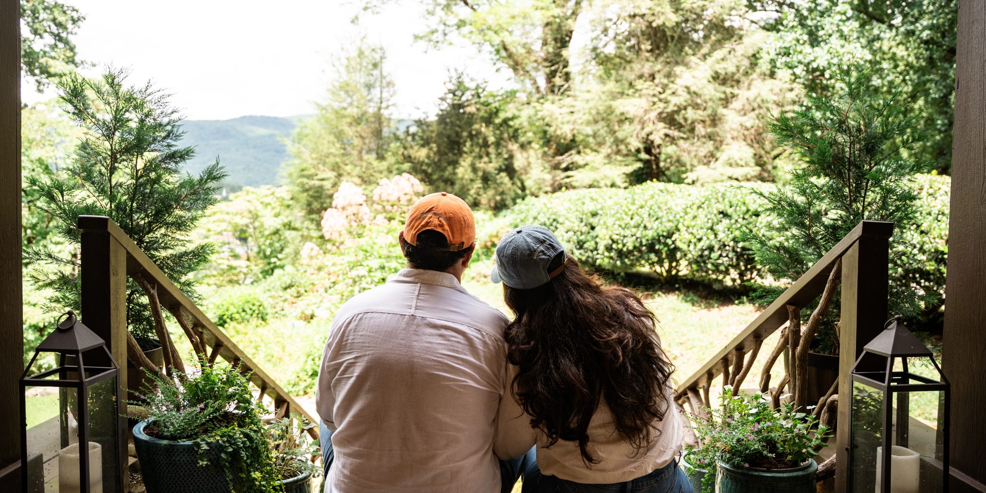 Couple sits on a wooden porch railing with their backs to the camera, looking out over a lush garden and hills beyond. An Authentic Bed and Breakfast Experience in the North Georgia Mountains