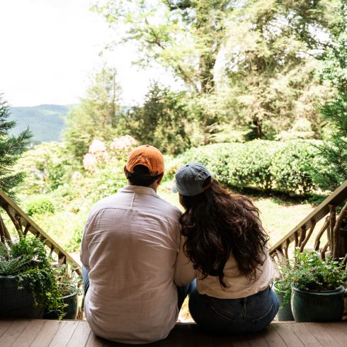 Couple sits on a wooden porch railing with their backs to the camera, looking out over a lush garden and hills beyond. An Authentic Bed and Breakfast Experience in the North Georgia Mountains