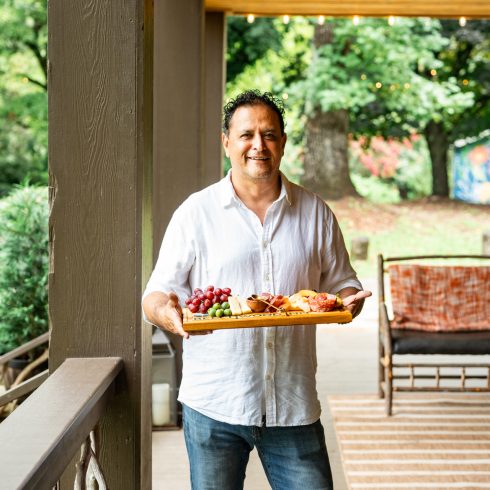 Smiling man in a white shirt holding a wooden board with grapes, cheese, and cured meats on a covered porch. An Authentic Bed and Breakfast Experience in the North Georgia Mountains