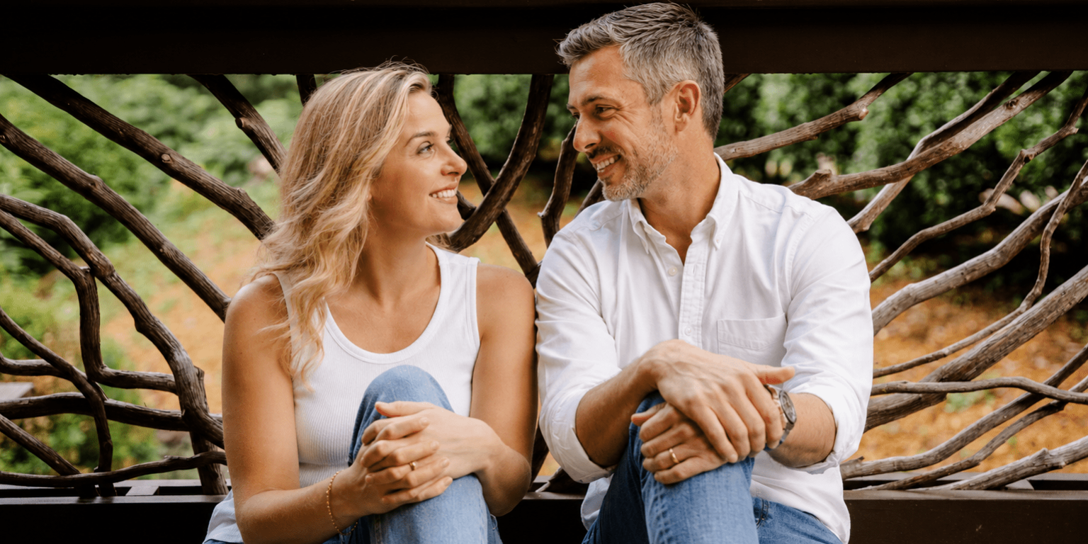 Smiling couple sitting close on a wooden bench outdoors, with intertwined branches behind them behind them.