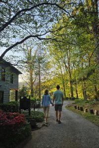 Couple walking hand in hand along a gravel path through a sunlit park with trees and a rustic house on the left.