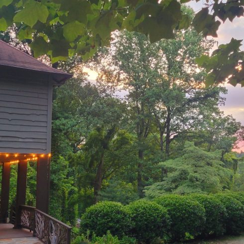 Side view of a gray wooden house with string lights on a covered porch, surrounded by lush trees at sunset.