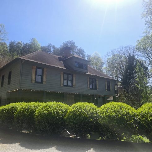Two-story green house with a brick chimney, surrounded by trees on a sunny day in a leafy yard.
