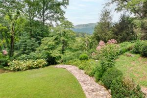 Stone walkway winds through a lush garden with flowering shrubs and distant hills under a blue sky.