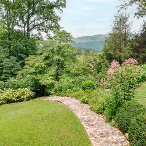 Stone walkway winds through a lush garden with flowering shrubs and distant hills under a blue sky.