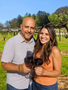 A smiling couple toasting with red wine glasses in a sunny vineyard, green grass and vines in the background.
