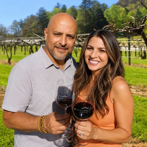 A smiling couple toasting with red wine glasses in a sunny vineyard, green grass and vines in the background.