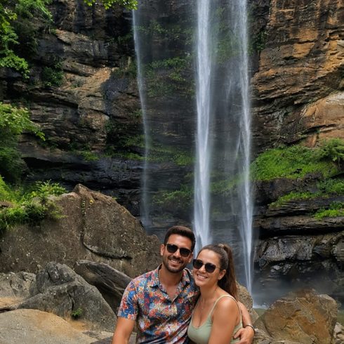 Couple sitting on rocks in front of a tall waterfall surrounded by green trees, smiling at the camera.