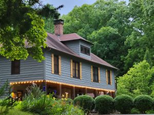 Gray two-story house with brown shutters, red roof, and brick chimney framed by lush green trees and garden foliage nearby.