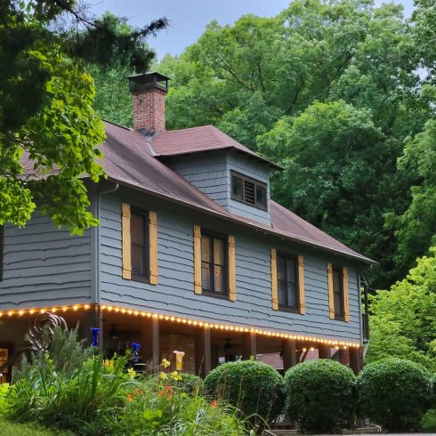 Gray two-story house with brown shutters, red roof, and brick chimney framed by lush green trees and garden foliage nearby.