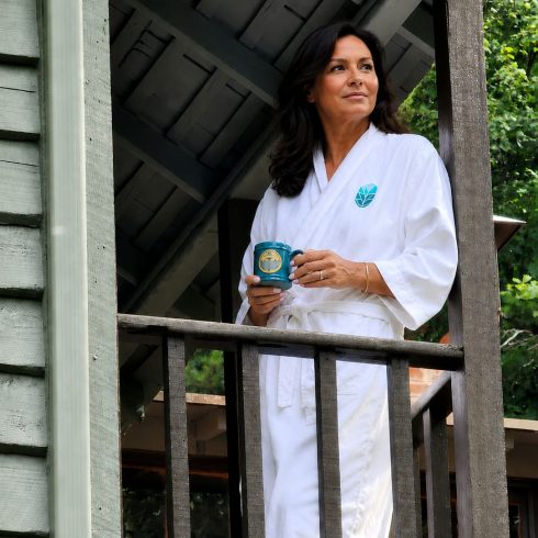 Woman in a white robe stands on a wooden balcony, holding a teal mug outdoors, with greenery in the background