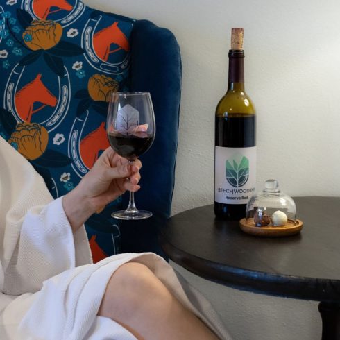 Person in a white robe holds a glass of red wine near a bottle on a side table with a dessert under a glass cloche
