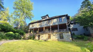 Two-story gray house with a stone base, wooden shutters, and a large wraparound porch surrounded by trees and a grassy yard.
