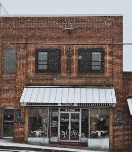 Snowy brick storefront with a metal awning, large display windows on the ground floor showing mannequins, and two upper-story windows; a quiet winter street scene.