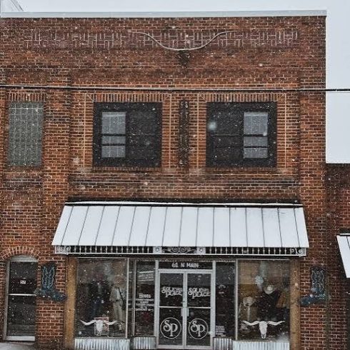 Snowy brick storefront in Clayton, GA, with a metal awning, large display windows on the ground floor showing mannequins, and two upper-story windows; a quiet winter street scene.