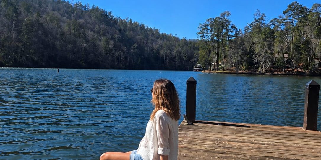 Person sitting on a wooden dock with feet in the lake, facing a forested lake under a clear blue sky.
