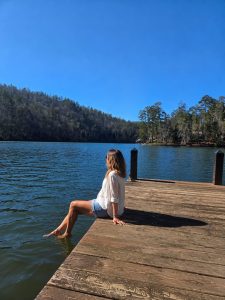 Person sitting on a wooden dock with feet in the lake, facing a forested lake under a clear blue sky.