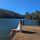 Person sitting on a wooden dock with feet in the lake, facing a forested lake under a clear blue sky.