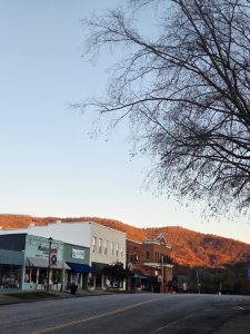 Street view of a small-town main street with storefronts, leafless tree branches overhead, and autumn hills in the distance.
