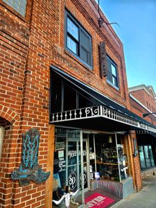 Brick two-story storefront with a black metal awning and a vertical sign reading'Sole Place' on the brick wall; blue metal sculpture mounted to the left and glass shopfront below with a red mat outside.
