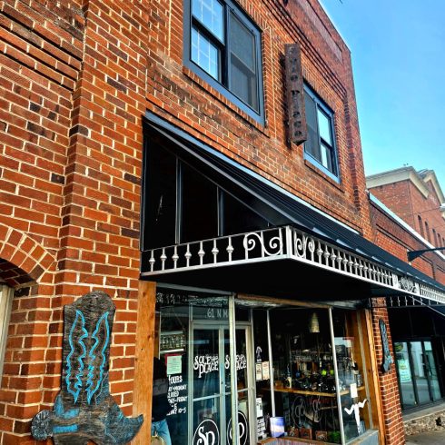 Brick two-story Sole Place storefront in Clayton, GA, with a black metal awning and a vertical sign reading “Sole Place” on the brick wall; blue metal sculpture mounted to the left and glass shopfront below with a red mat outside.