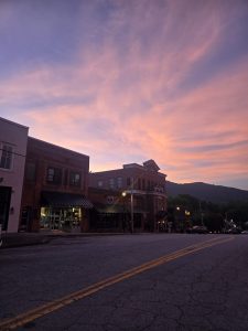 Downtown street at dusk with pink and purple sky, brick buildings, and mountain silhouette in the background.
