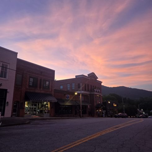 Downtown Clayton, GA street at dusk with pink and purple sky, brick buildings, and mountain silhouette in the background.