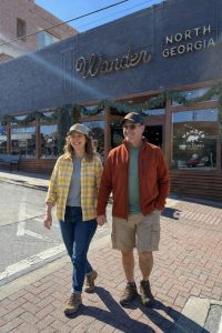 A smiling couple walks hand in hand on a sunlit street in front of a storefront that reads'Wander North Georgia'. The woman wears a yellow plaid shirt and cap, the man a rust-colored jacket and cap.