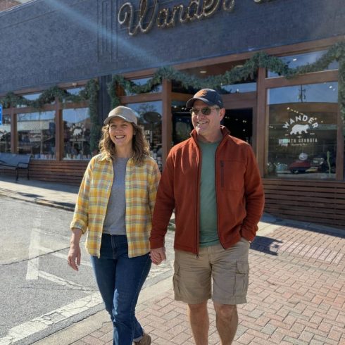 A smiling couple walks hand in hand on a sunlit street in Clayton, GA, in front of a storefront that reads “Wander North Georgia.” The woman wears a yellow plaid shirt and cap, the man a rust-colored jacket and cap.