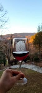 Hand holding a wine glass with red wine outdoors at sunset, with rolling hills and trees in the background, glass shows a leaf logo.
