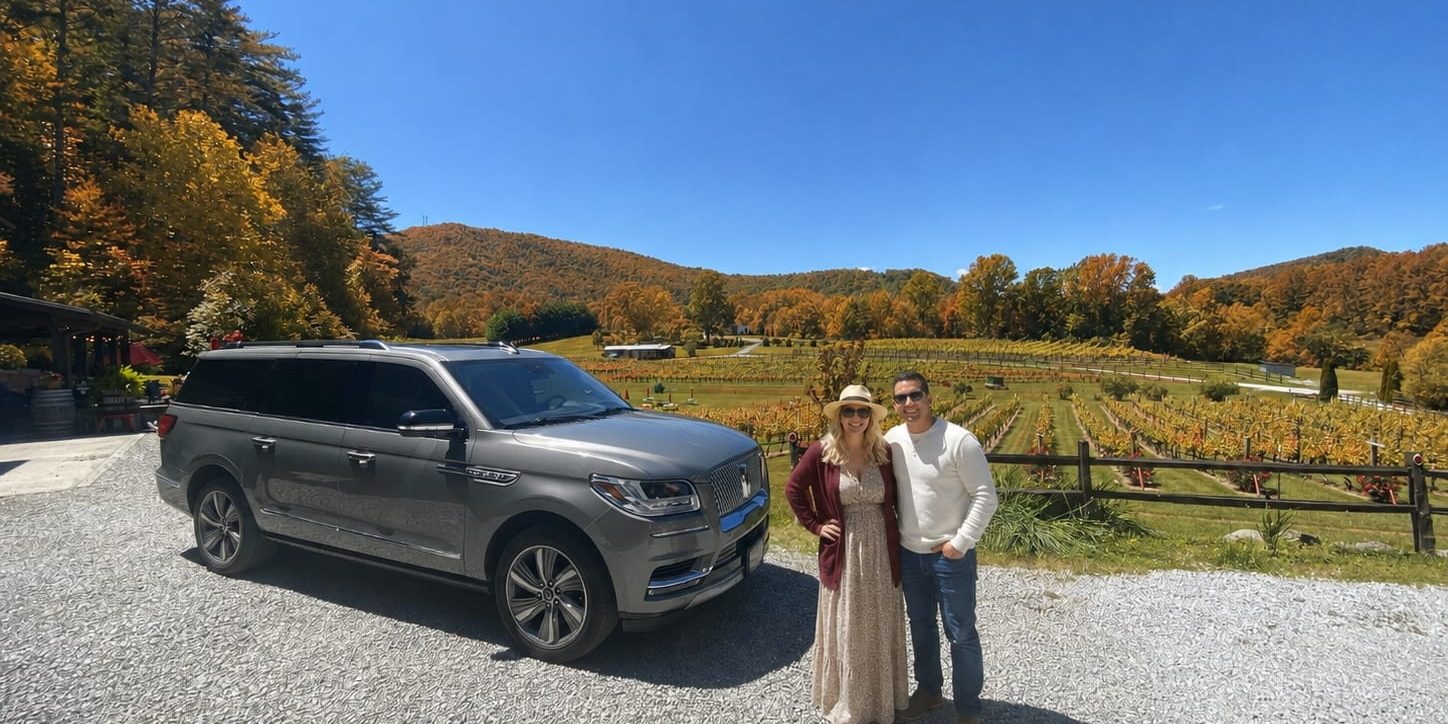 Gray SUV parked on gravel beside a vineyard; a smiling couple in autumn clothes pose in front of colorful fall trees under a blue sky.