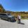 Gray SUV parked on gravel beside a vineyard; a smiling couple in autumn clothes pose in front of colorful fall trees under a blue sky.