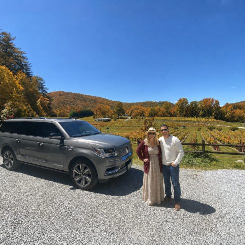 Gray SUV parked on gravel beside a vineyard; a smiling couple in autumn clothes pose in front of colorful fall trees under a blue sky.