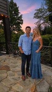 Couple standing on a stone patio at sunset, arms around each other, wearing blue denim outfits and smiling.