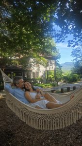 Couple relaxing in a white woven hammock outdoors beside a wooden house, green trees overhead and a mountain view in the background, smiling and cuddling.