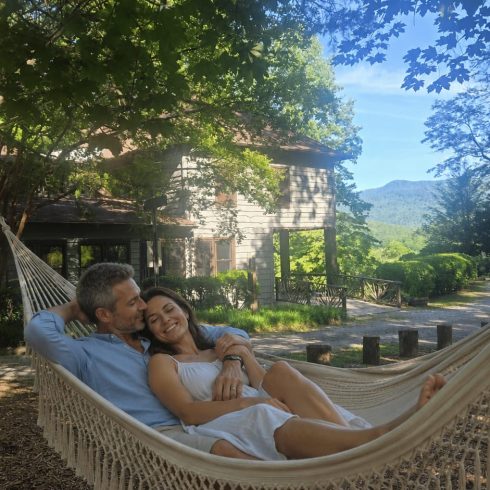 Couple relaxing in a white woven hammock outdoors beside a wooden house, green trees overhead and a mountain view in the background, smiling and cuddling.