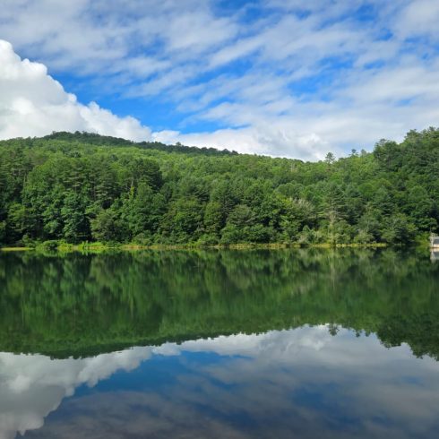 Calm lake reflecting the green forest and blue sky with white clouds; a small dock on the right shore.