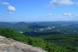 Wide view over green forested mountains and a distant valley town under a blue sky with scattered clouds.