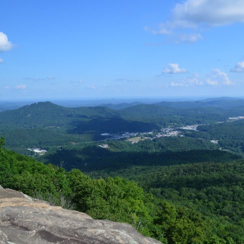 Wide view over green forested mountains and a distant valley town under a blue sky with scattered clouds.