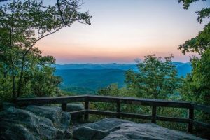 Sunset over layered blue mountain ridges viewed from a rocky overlook with a wooden railing.