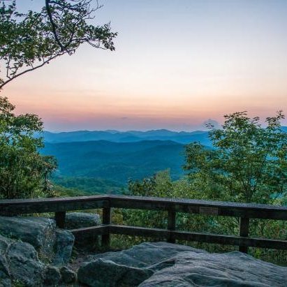 Sunset over layered blue mountain ridges viewed from a rocky overlook with a wooden railing.
