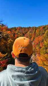 Back view of a person wearing an orange cap and light blue hoodie, overlooking a colorful autumn forest under a clear blue sky.