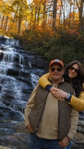 Couple posing arm-in-arm in front of a multi-tiered waterfall with autumn trees in the background.