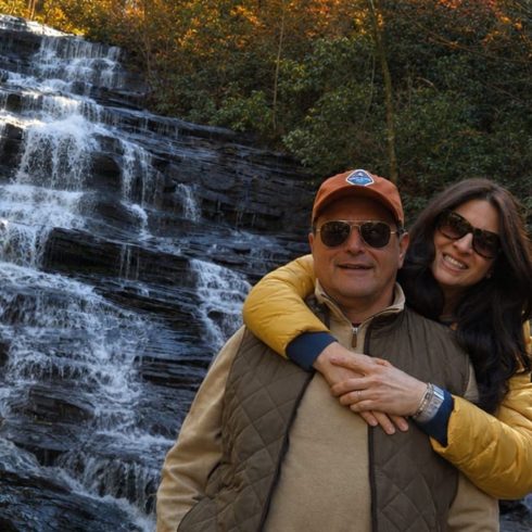 Couple posing arm-in-arm in front of a multi-tiered waterfall with autumn trees in the background.