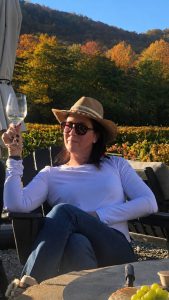 Woman in a white long-sleeve shirt and straw hat sits in a chair, holding a wine glass, with an autumn vineyard and hills behind her.
