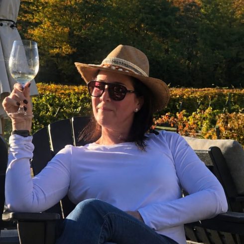 Woman in a white long-sleeve shirt and straw hat sits in a chair, holding a wine glass, with an autumn vineyard and hills behind her.