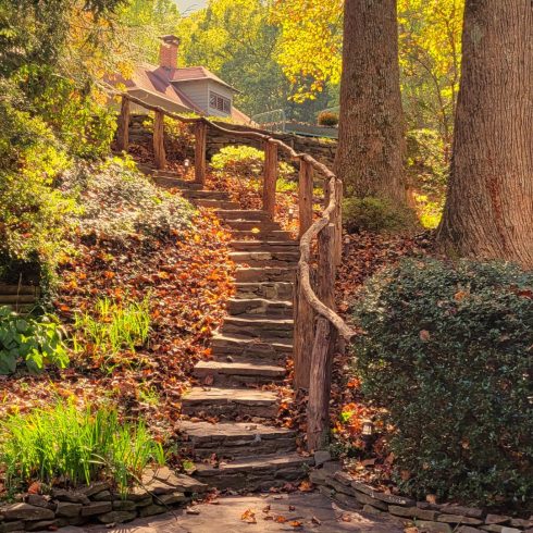 Autumn garden scene with orange pumpkins at the front, stone steps and a rustic wooden railing winding up a slope toward a cottage.