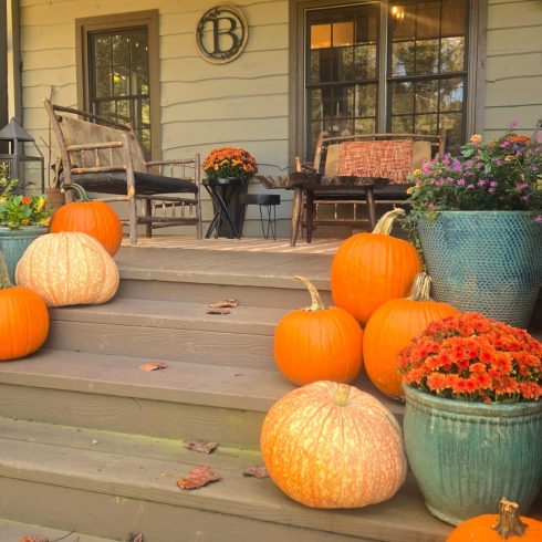 Front porch decorated for autumn with pumpkins, mums, and planters on wooden steps; cozy seating under a wooden ceiling.