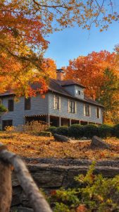 Blue two-story wooden house with a wraparound porch, string lights along the edge, surrounded by vibrant orange autumn leaves and a clear blue sky.