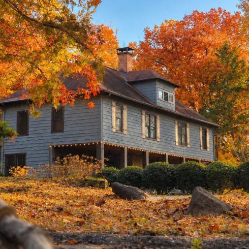 Blue two-story wooden house with a wraparound porch, string lights along the edge, surrounded by vibrant orange autumn leaves and a clear blue sky.